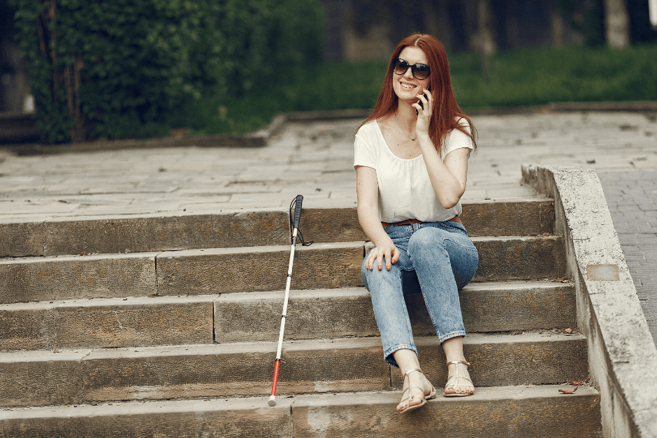 A feminine-presenting individual using her phone on some steps with her cane next to her.