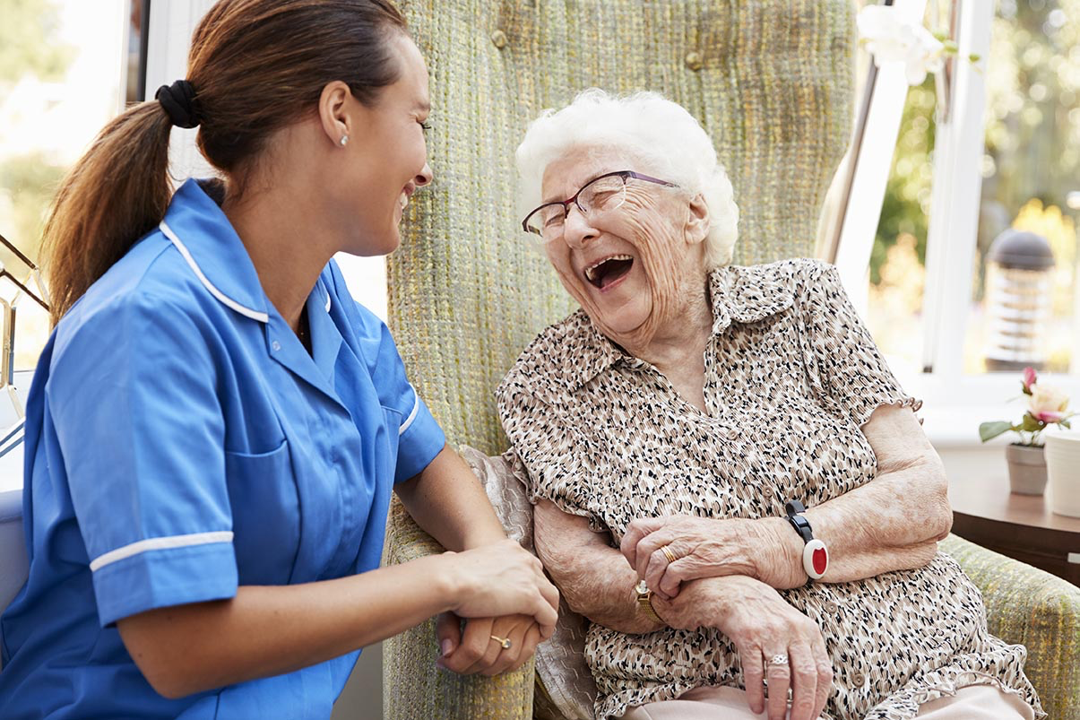 A female brunette caregiver wearing a blue uniform top talking to an elderly woman with white hair and leopard spots patterned button up shirt.