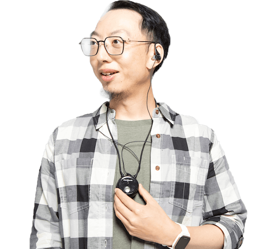 An Asian man with black-rimmed glasses and hair, long sleeve plaid shirt and a green t-shirt, listening through a device and smiling.