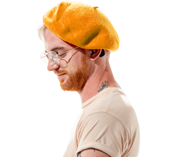 A white Deaf man wearing a gold hat and gold-rimmed glasses. The man is wearing a tan t-shirt and has a flower tattoo on his left neck.