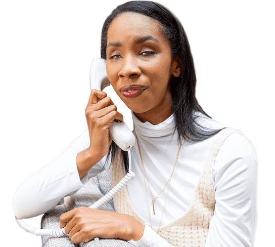 A blind Black woman wearing a white turtleneck shirt and blouse sitting on the couch, holding a white phone to her ear, listening to the caller on the other end of the line.